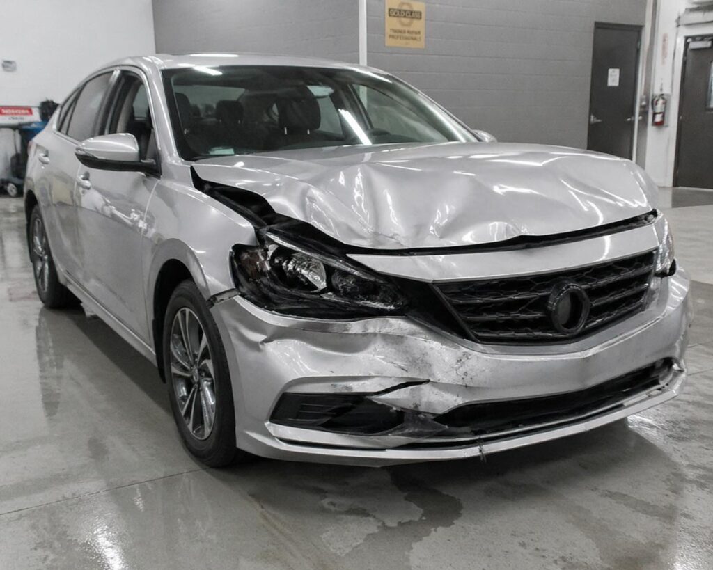 Sedan with crumpled front-end collision damage including hood, bumper, and headlight in body shop intake bay.